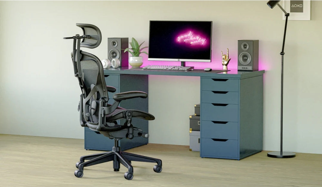 A modern home office with a Refurne Atlas Headrest for Herman Miller Aeron Remastered Chair (Graphite) for ergonomic support, blue-drawer desk, monitor with pink neon text, speakers, keyboard, plant, gold hand decor—all on light wood flooring.