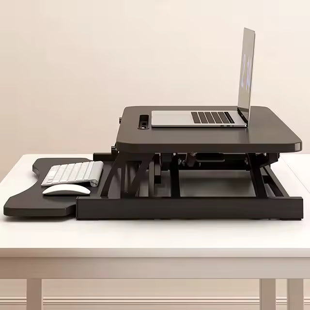 The Refurne Height Adjustable Standing Desk Riser Table creates an ergonomic setup, with a laptop on top and a wireless keyboard and mouse on its pull-out tray, arranged on a white desk against a beige wall.