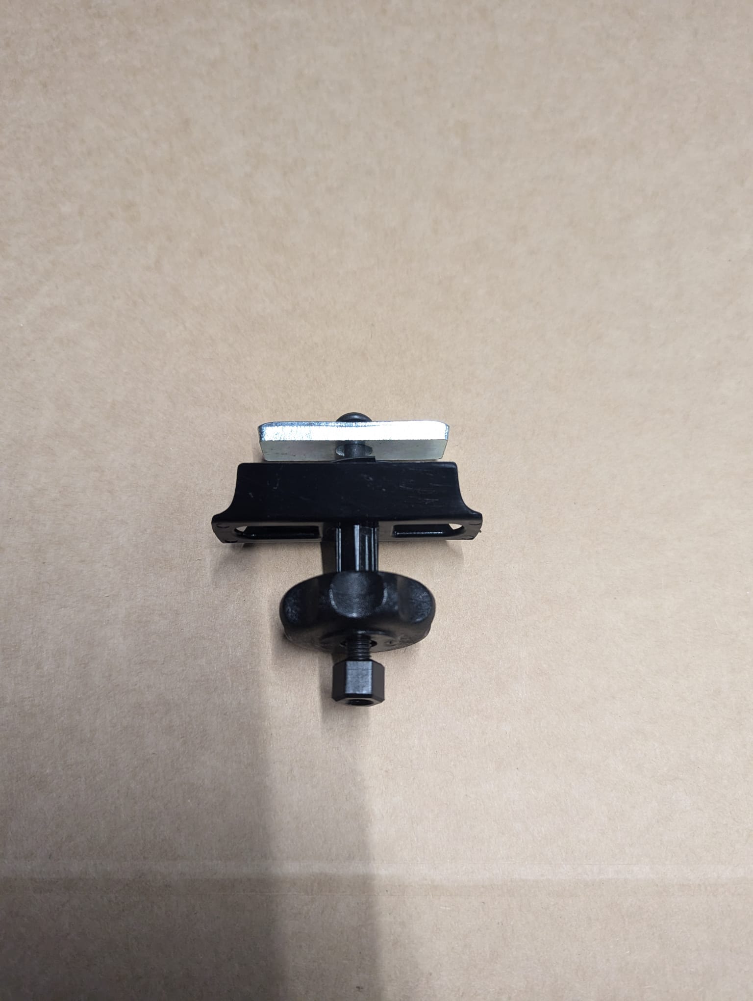 A black metal clamp with a threaded knob and silver plate from Refurne, the Roller/Wheel Arm Mechanism for Herman Miller Classic Aeron Chair MK1, is shown on a plain light brown surface.